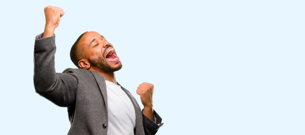 Африканский американец с бородой счастлив и взволнован, празднуя победу, выражая большой успех, силу, энергию и положительные эмоции. Празднует новую работу радостно изолированы на голубом фоне
 - Фото, изображение