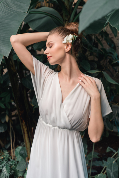 attractive tender bride in white dress with flower in hair posing in tropical garden - 写真・画像