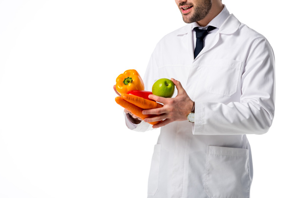 cropped shot of nutritionist in white coat with fresh vegetables and apple in hands isolated on white - Фото, зображення