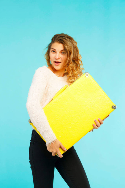 Young happy beautiful woman holding yellow suitcase over blue ba - Foto, Imagen
