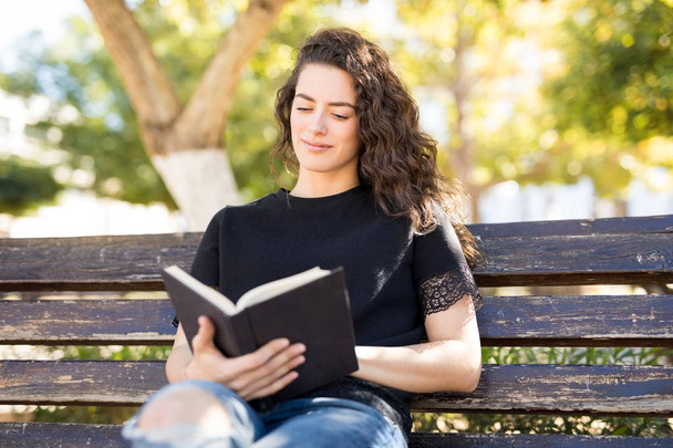 wunderschöne Frau in ihren Zwanzigern, die es sich auf einer Parkbank gemütlich macht und ein Buch liest - Foto, Bild