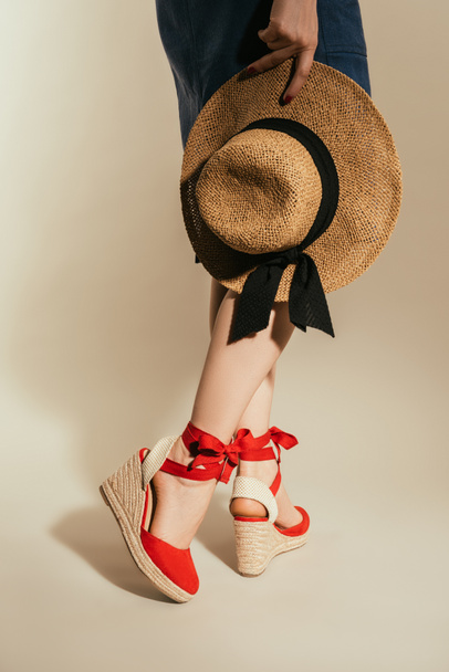 corte tiro de mulher elegante em sandálias plataforma vermelha segurando chapéu de palha no fundo bege
 - Foto, Imagem