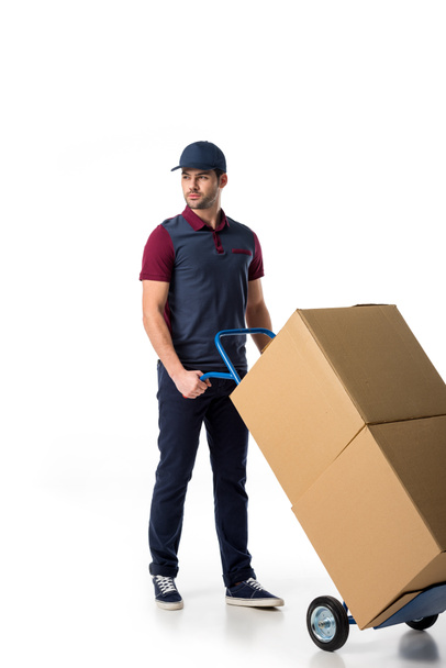 delivery man in uniform pushing hand truck with cardboard boxes isolated on white - Foto, Imagen
