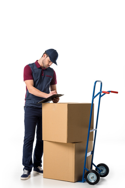 delivery man in uniform checking notes in notepad with cardboard boxes on hand truck near by isolated on white - Foto, Imagen