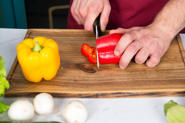 Hand slice pepper with ceramic knife. Vegetables getting cut on wooden cutting board. Food preparation and cooking recipes. Vegetarian menu and healthy diet. Organic products concept - Photo, image