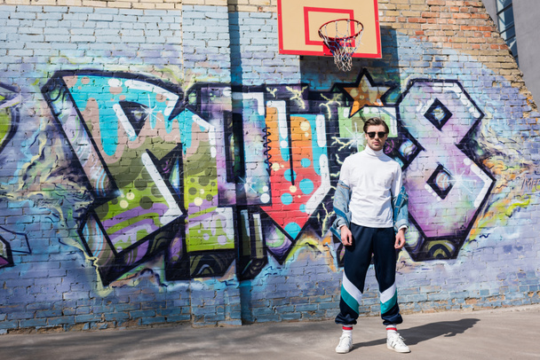 stijlvolle jongeman in vintage kleding voor bakstenen muur met graffiti en basketbal ring - Foto, afbeelding