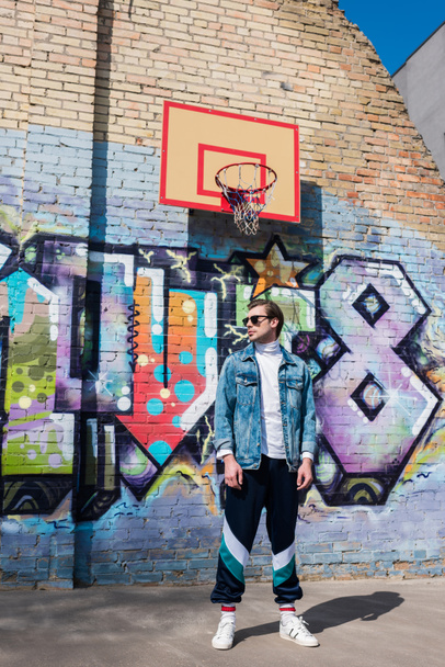 knappe jongeman in vintage kleding voor bakstenen muur met graffiti en basketbal ring - Foto, afbeelding