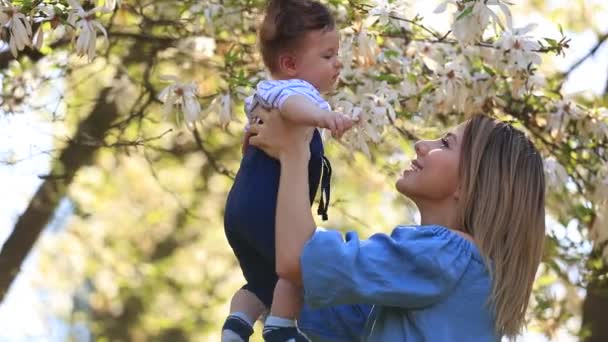 Jonge roodharige moeder en kind hebben een vrijetijdsbesteding in lente magnolia bloeiende tuin op zonnige dag - Video