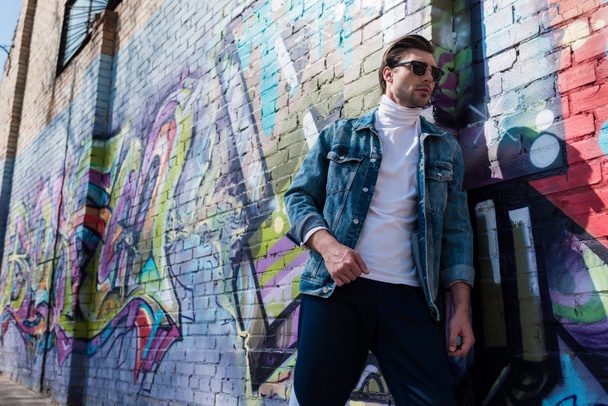 stylish young man in vintage clothing leaning on brick wall with graffiti - Фото, зображення