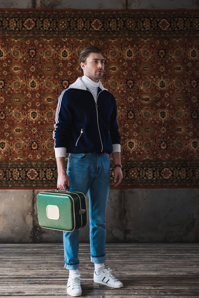 handsome man with vintage little suitcase in front of rug hanging on wall - Photo, Image