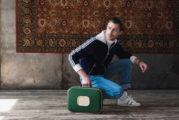 joven guapo con la pequeña maleta vintage sentado sentada en cuclillas delante de la alfombra colgando en la pared
 - Foto, imagen