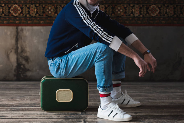 cropped shot of man sitting on vintage little suitcase in front of rug hanging on wall - Foto, afbeelding