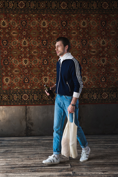 jeune homme en vieux vêtements scolaires avec un sac à ficelle et une bouteille de bière devant un tapis accroché au mur
 - Photo, image