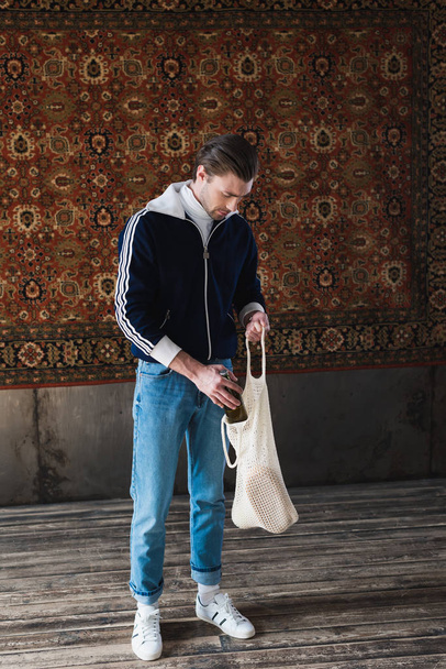 man in old school clothes putting bottle of beer into string bag in front of rug hanging on wall - Photo, Image