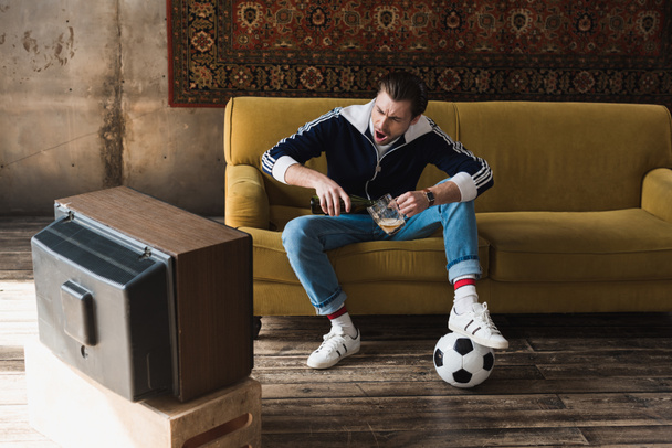schreiender junger Mann in Vintage-Klamotten mit Ball beim Fußballgucken im alten Fernseher und dem Einschenken von Bier in Krug - Foto, Bild