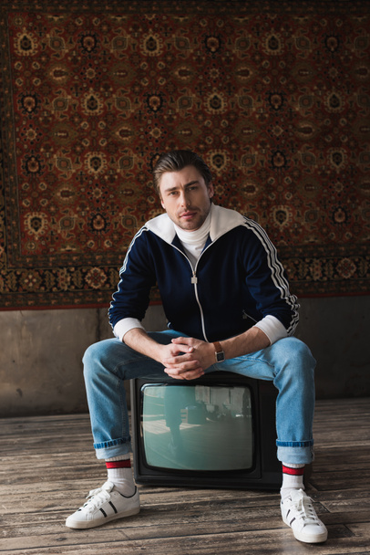 handsome young man in vintage clothes sitting on retro tv set and looking at camera in front of rug hanging on wall - Fotografie, Obrázek