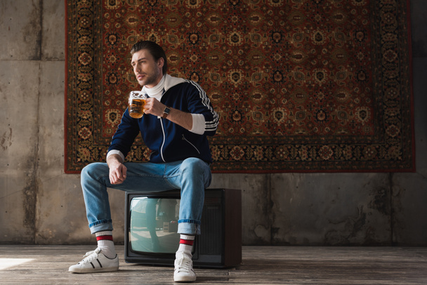 Nachdenklicher junger Mann in Vintage-Kleidung mit einem Becher Bier sitzt auf einem Retro-Fernseher vor einem Teppich, der an der Wand hängt - Foto, Bild