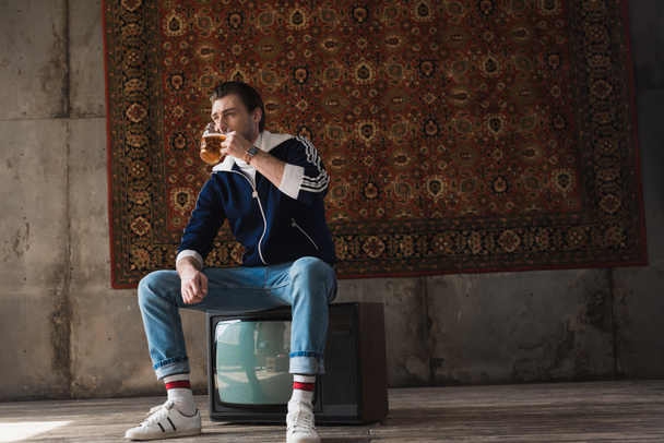 handsome young man in vintage clothes drinking beer while sitting on retro tv set in front of rug hanging on wall - Photo, image