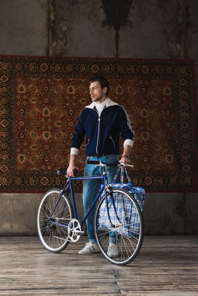 handsome young man in vintage clothes with road bike and bag in front of rug hanging on wall - Fotografie, Obrázek