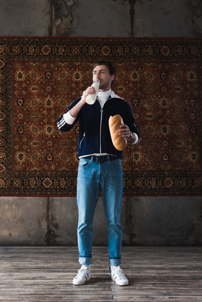 joven guapo en ropa vintage con botella de leche y pan delante de la alfombra colgando en la pared
 - Foto, Imagen