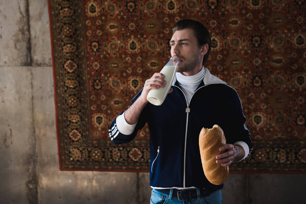 νεαρός άνδρας στο vintage ρούχα με μπουκάλι γάλα και καρβέλι ψωμί μπροστά από την κουβέρτα που κρέμονται στον τοίχο - Φωτογραφία, εικόνα