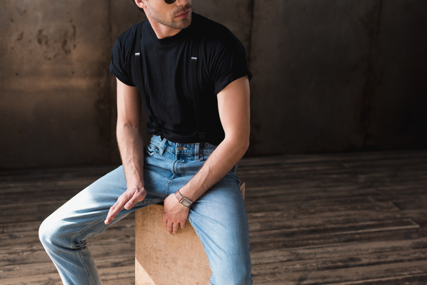 cropped shot of young man in black t-shirt and suspenders sitting on wooden box - Фото, зображення
