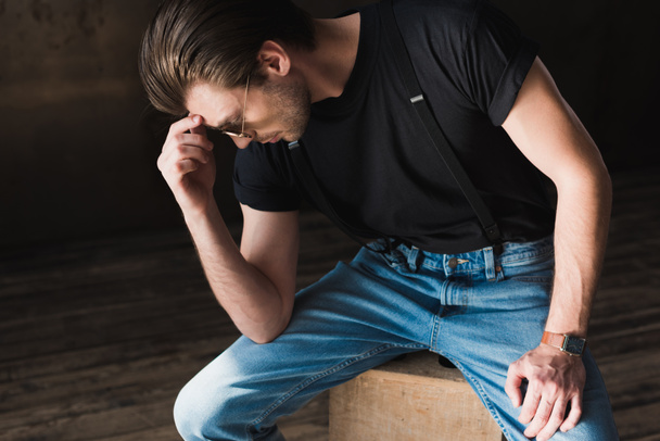thoughtful young man in black t-shirt and suspenders sitting on wooden box - Фото, зображення