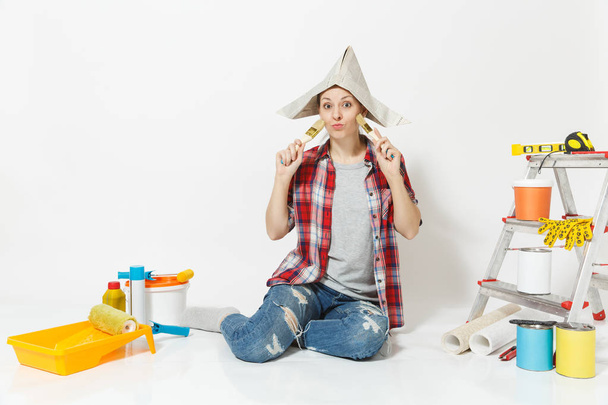 Διασκεδαστικό γυναίκα στην εφημερίδα καπέλο κάθεται στο πάτωμα με πινέλο, όργανα για ανακαίνιση Διαμέρισμα Δωμάτιο απομονωθεί σε λευκό φόντο. Ταπετσαρία, αξεσουάρ για την κόλληση, εργαλεία ζωγραφικής. Επισκευή concept home. - Φωτογραφία, εικόνα