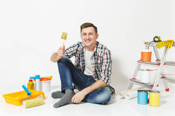 lächelnder lustiger Mann, der mit Pinsel auf dem Boden sitzt, Instrumente für die Renovierung seines Wohnzimmers isoliert auf weißem Hintergrund. Tapeten, Klebezubehör, Malwerkzeuge. Heimkonzept reparieren. - Foto, Bild