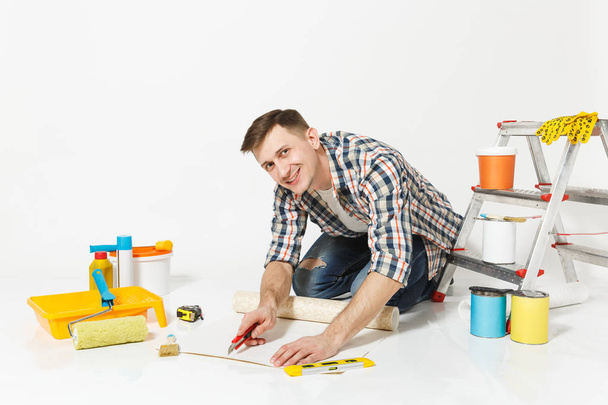 Man sitting on floor with wallpaper roll, stationery knife, instruments for renovation apartment room isolated on white background. Wallpaper, gluing accessories, painting tools. Repair home concept. - Fotografie, Obrázek