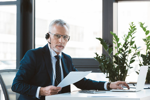 serious businessman in eyeglasses and headset using laptop and looking at camera - Fotoğraf, Görsel