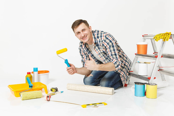 Young man sitting on floor with wallpaper roll, paint roller, instruments for renovation apartment room isolated on white background. Wallpaper, gluing accessories, painting tools. Repair home concept - Photo, Image
