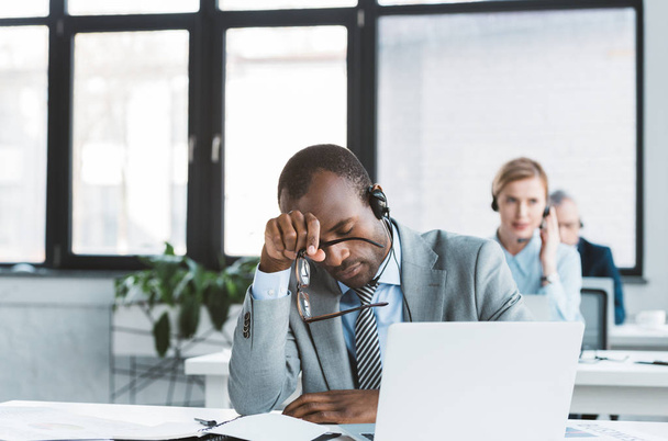 hombre de negocios afroamericano cansado en auriculares con gafas mientras trabaja con el ordenador portátil en la oficina
 - Foto, Imagen