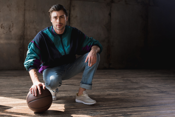 attractive young man in vintage windcheater with basketball ball sitting squats on wooden floor - Valokuva, kuva