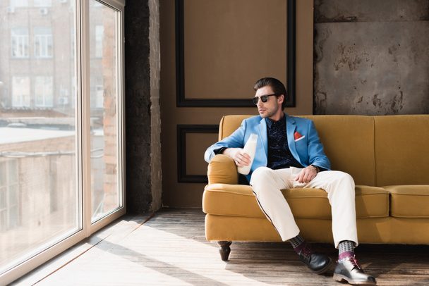 atraktivní mladý muž v elegantní oblek sedí na gauči s lahví mléka - Fotografie, Obrázek