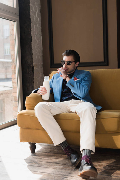 attractive young man in stylish suit sitting on couch with bottle of milk and wiping mouth with hand - Foto, imagen