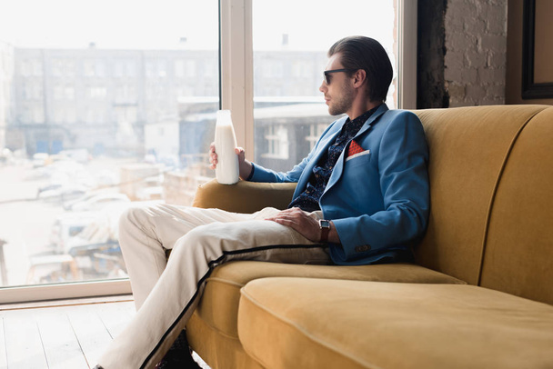 thoughtful young man in stylish suit sitting on couch with bottle of milk and looking through window - Foto, Imagen