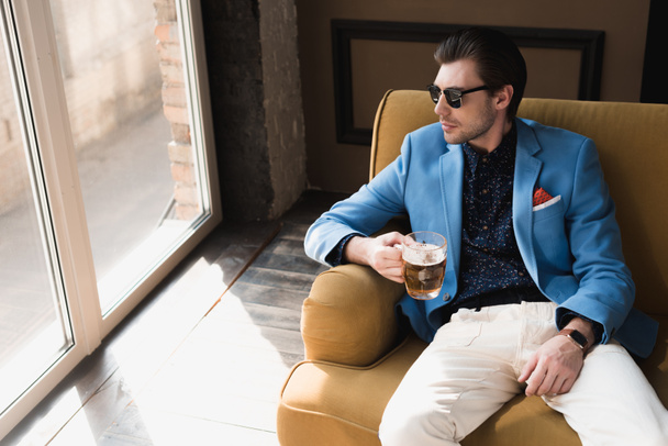 vue grand angle de attrayant jeune homme en costume élégant assis sur le canapé avec tasse de bière
 - Photo, image