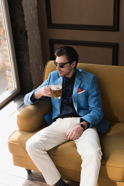 high angle view of handsome young man in stylish suit sitting on couch with mug of beer - Фото, зображення