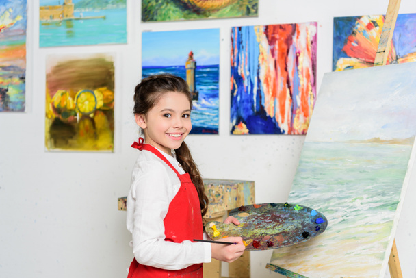 preteen kid holding palette near canvas in workshop of art school - Photo, Image