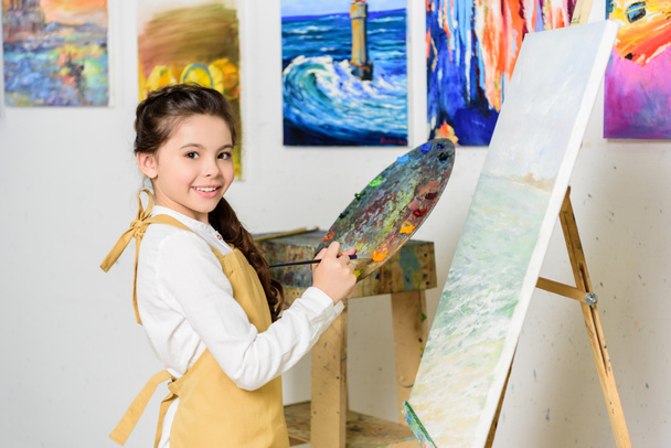 Kind hält Palette in der Hand und blickt in die Kamera in der Werkstatt der Kunstschule - Foto, Bild