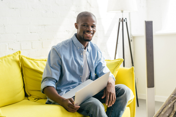 portret uśmiechający się Afro-Amerykanin z laptopa siedząc na kanapie w domu - Zdjęcie, obraz