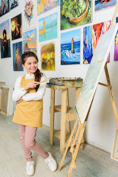 smiling kid standing with painting brush near canvas in workshop of art school - Foto, Imagen