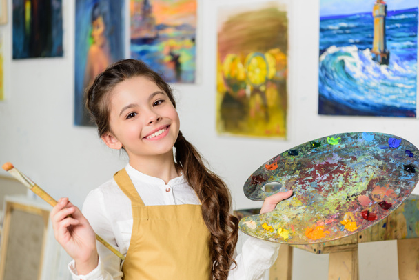 Lächelndes Kind steht mit Palette und Pinsel in der Werkstatt der Kunstschule - Foto, Bild