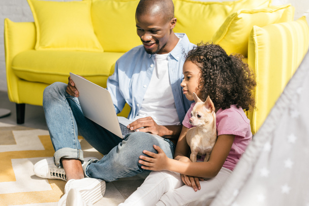 Afrikaanse Amerikaanse vader en dochtertje met hond met behulp van laptop samen thuis - Foto, afbeelding