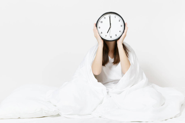 顔、白いシーツ、枕、白い背景の上の毛布に包む丸い時計の前でベッドに座っている女性。女性目を覚ます朝、部屋で時間を過ごします。残りの部分がリラックスして、良い気分のコンセプト - 写真・画像
