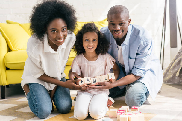 african american parents and daughter holding wooden blocks with family lettering together on floor at home - Fotografie, Obrázek