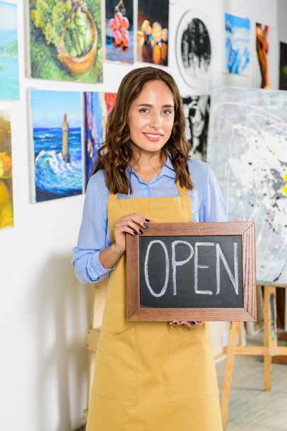 attractive female artist holding signboard with word open in workshop - Photo, Image