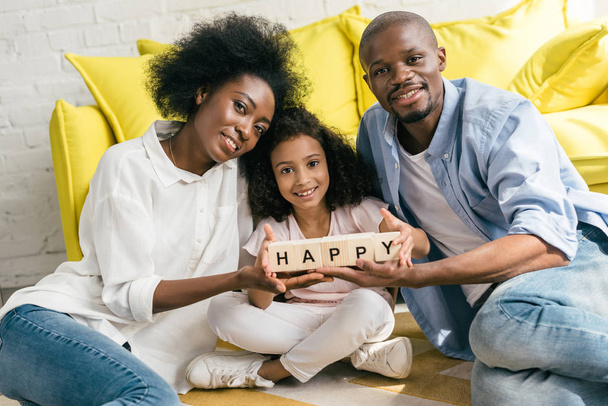 afrikanisch-amerikanische Eltern und Tochter halten zu Hause Holzklötze mit fröhlichen Schriftzügen zusammen auf dem Boden - Foto, Bild