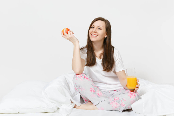 Young brunette woman sits on bed with white sheet, pillow, wrapping in blanket isolated on white background. Beauty female have breakfast with apple, fresh orange juice. Rest, relax, good mood concept - Foto, Imagem
